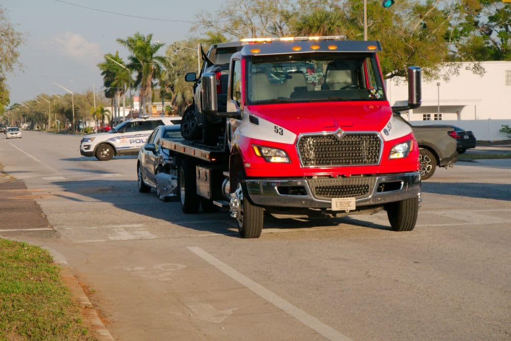 Car accident after semi truck collision – Personal Injury Lawyer in Fort Lauderdale Tow truck with damaged car at accident scene on a city street, police vehicle nearby under clear skies.