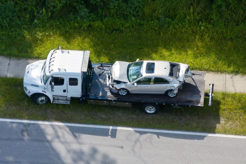 commercial trucking accident – Personal Injury Lawyer in Fort Lauderdale Tow truck transporting a damaged silver car on a sunny roadside, viewed from above.